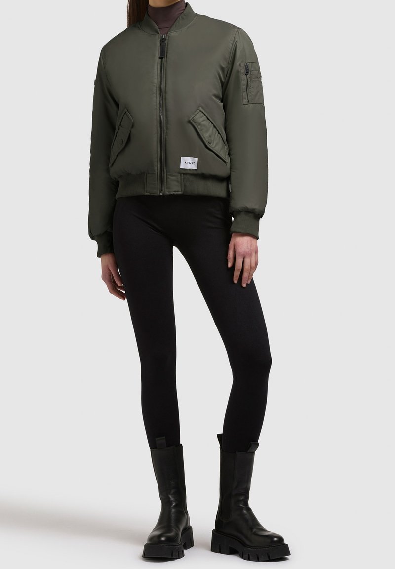 Personne portant une veste bomber verte, des leggings noirs et des bottes noires épaisses, debout devant un fond uni.