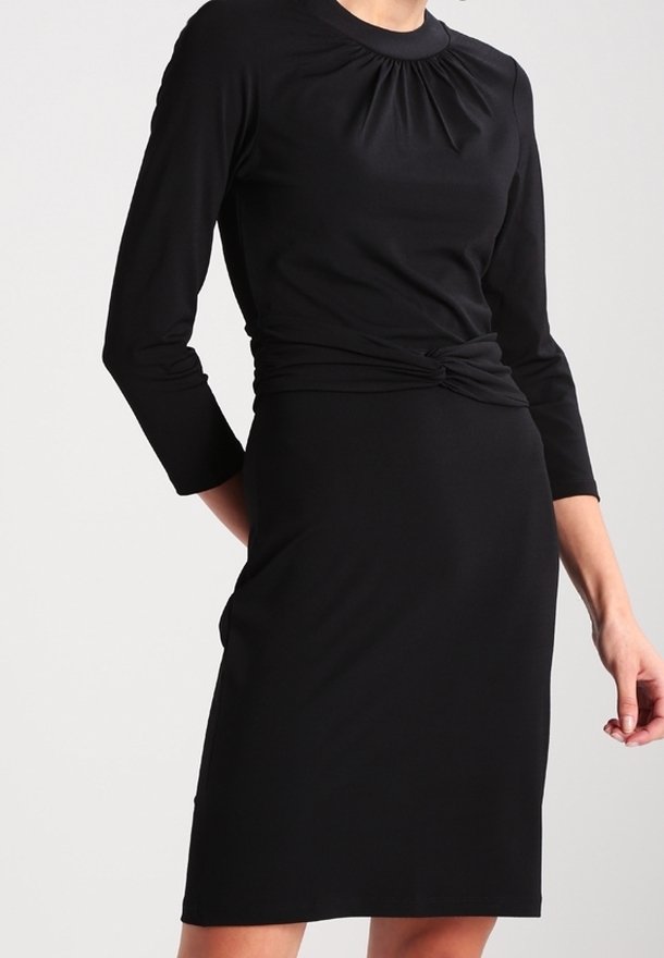Femme portant une robe noire ajustée à manches trois-quarts, avec un col plissé et un détail noué à la taille.