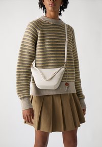 Helle beigefarbene Umhängetasche mit einer geschwungenen Form und einem strukturierten Klappenverschluss, kombiniert mit einem gestreiften Pullover und einem plissierten khakifarbenen Rock.