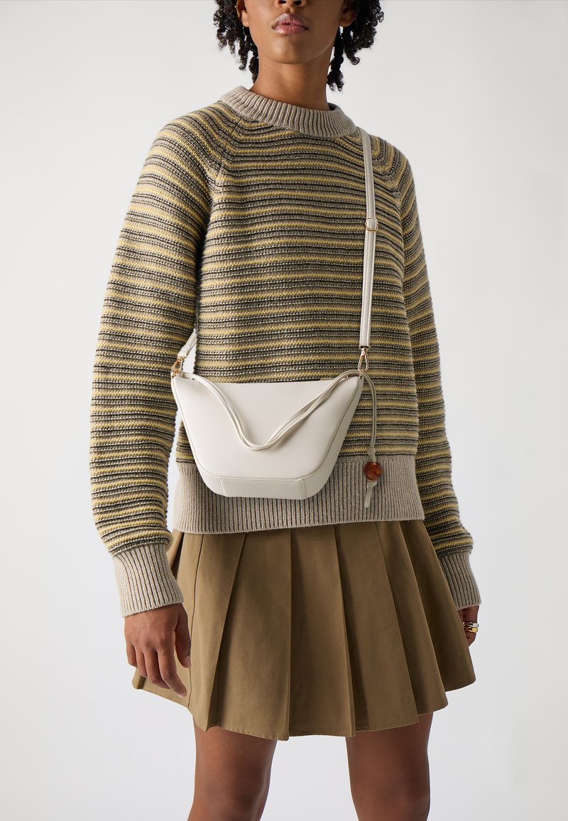 Kerge beez crossbody kott, millel on kaarjas disain ja tekstuuriga klapp, sobituna triibulise sviitriga ja volditud khaki seelikuga.