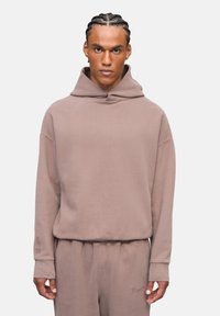 Uomo con capelli intrecciati che indossa un hoodie taupe e pantaloni della tuta coordinati, in piedi contro uno sfondo grigio chiaro uniforme.