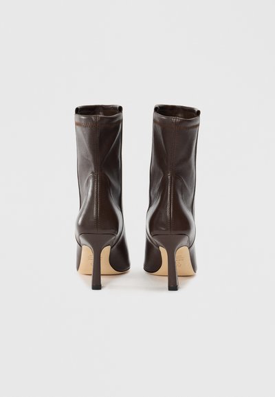 Un paio di stivaletti in pelle marrone scuro con alti tacchi a stiletto, visti da dietro su uno sfondo bianco semplice.