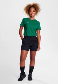 Camiseta verde de manga corta con logo, pantalones cortos con cintura elástica negra, calcetas altas negras y zapatillas deportivas negras en un modelo.