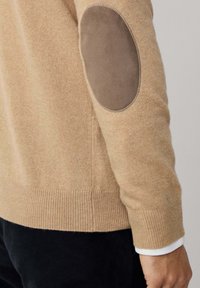 Bežani pleteni pulover s smešnim smešnim kožnim krpicama na laktovima, nošen preko belih rukava, uparen s crnim hlačama.