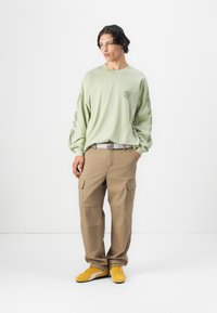 Chemise à manches longues vert clair avec un logo sur le devant et un design graphique sur les manches, assortie à un pantalon cargo beige et des baskets jaunes.