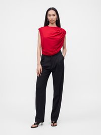 Femme debout portant un haut rouge sans manches drapé, un pantalon noir taille haute, des sandales noires à talons et des boucles d'oreilles créoles dorées sur un fond blanc.