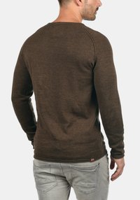 Rjav dolg rokav pulover z teksturirano pletenino, okroglo izrezanega vratu in prilegajočimi rokavi. Ima majhno oznako na robu. Nosi se z svetlo sivo jeans hlačami.