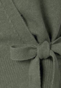 Cardigan lavorato a maglia in verde oliva, con una superficie testurizzata e una cintura di stoffa legata in vita, con un motivo a costine visibile sui bordi.