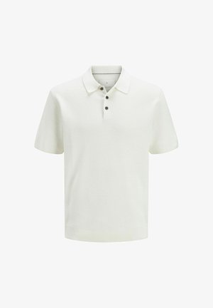 Niño con pelo rizado de pie, vistiendo una camiseta polo blanca de manga corta, pantalones cortos negros, calcetines blancos y zapatillas grises sobre un fondo blanco.