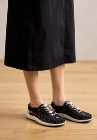 Black mesh athletic shoes with a crisscross design, white laces, and a cushioned white sole; open structure for ventilation.