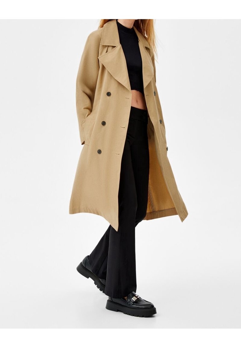 Femme marchant avec un trench-coat beige croisé, un top court noir, un pantalon large noir et des mocassins noirs sur fond blanc.