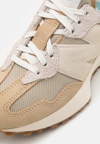Beżowy but sportowy z siateczką i zamszowymi materiałami, wyposażony w białe sznurowadła, teksturowane nakładki oraz gumową podeszwę z wzorem zapewniającym przyczepność.