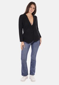 Blazer nero con scollatura a V, chiusura a un bottone e maniche lunghe, abbinato a jeans blu e sneaker bianche.