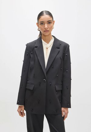 Mujer con el cabello oscuro recogido lleva una chaqueta negra adornada con pequeños racimos de cuentas negras sobre una blusa blanca, de pie contra un fondo liso.