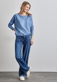 Maglione azzurro chiaro con una vestibilità rilassata, maniche lunghe; abbinato a jeans a gamba larga in denim scuro e sneakers bianche, in piedi contro uno sfondo neutro.