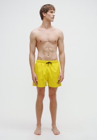 Dzelteni peldēšanas šorti ar regulējamu auklu jostasvietā, strukturēta auduma, virs ceļa garuma, ar zīmola logotipu uz kreisās kājas. Modelis stāv basām kājām.