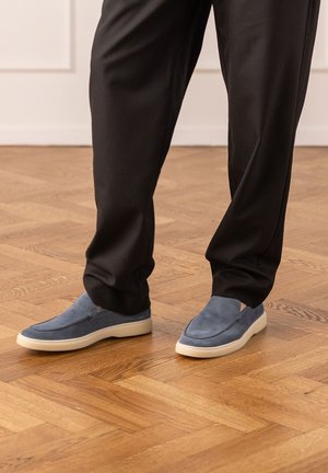 Uomo con pantaloni scuri e scarpe slip-on blu in piedi su un pavimento in parquet di legno all'interno.