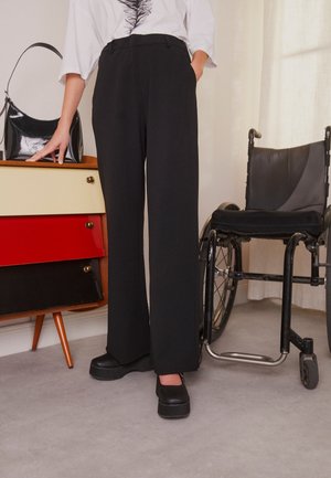 Osoba ubrana w czarne spodnie z wysokim stanem i buty na platformie stoi obok czarnego wózka inwalidzkiego oraz małej kolorowej szafki z czarną torebką.