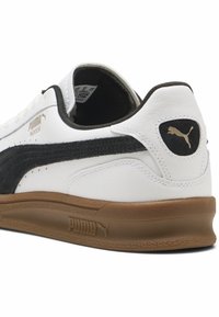 Baskets d'intérieur Puma blanches et noires avec semelle marron, montrant les logos latéraux et arrière ainsi que les détails des coutures sur fond blanc.
