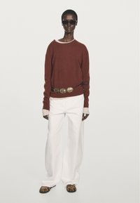 Roter Strickpullover mit weißem Kragen, weißen weit geschnittenen Hosen und einem dekorativen goldenen Gürtel. Das Model trägt schwarze Sonnenbrillen und gemusterte Sandalen.