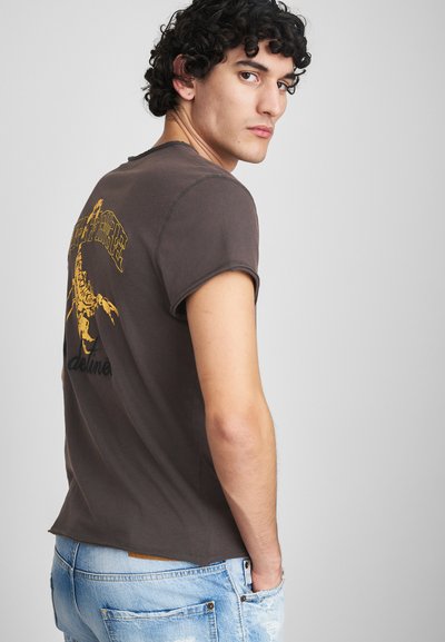 T-shirt di cotone marrone con collo rotondo, maniche corte e una grafica di scorpione gialla sul retro, abbinato a jeans chiari strappati.