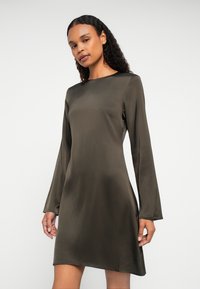 Sieviete ar piena matiem, tērpusies garās piedurknēs olīvzaļā satīna kleitā, stāv uz neitrāla fona.