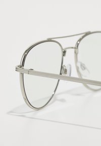 Lunettes de soleil en métal argenté avec des verres ronds, dotées de branches fines, de coussinets de nez transparents et d'un design minimaliste. Les verres semblent légèrement teintés.