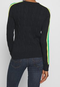 Marinblå kabelstickad tröja med ribbad nederkant, prydd med livfulla gröna och gula ränder längs axelsömmarna. Bärs tillsammans med mörka jeans.