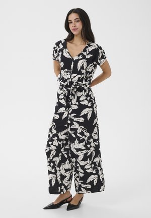 Femme debout, les mains derrière le dos, portant une combinaison pantalon noire à jambes larges avec des motifs de feuilles blanches et des chaussures slingback noires à bouts pointus.