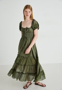 Olijfgroene maxi-jurk met korte pofmouwen, smock-borstige, gelaagde kanten zoom en een vloeiende ontwerp. Gepaard met zwarte platte sandalen.