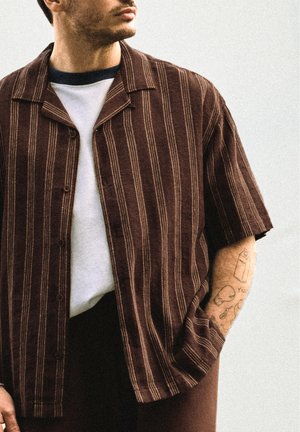 Homme portant une chemise marron à rayures verticales à manches courtes par-dessus un T-shirt blanc, avec la main dans la poche montrant des tatouages sur l’avant-bras, y compris un carton et un crâne.
