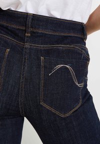Jeans de mezclilla oscura con un ajuste ceñido, que presentan costuras amarillas, un diseño distintivo en el bolsillo trasero y una textura suave.