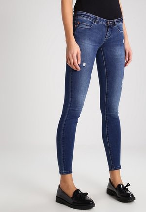 Slim-fit blauwe spijkerbroek met lichte slijtage, gedragen met zwarte loafers met franjes en een zwart topje dat zichtbaar is bij de taille.