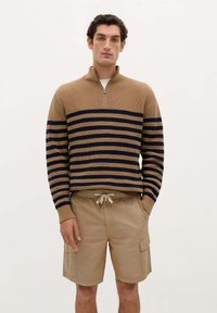 Brun og marineblå stribet sweater med høj krave og lynlås, parret med beige cargo shorts med sidelommer og elastisk talje med snor.