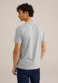 Camiseta gris de manga corta hecha de algodón, con cuello redondo y una textura suave. Lleva puesta una persona de espaldas, combinada con jeans oscuros.