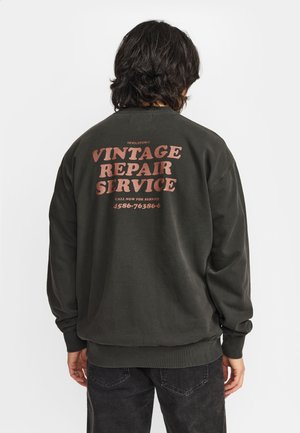 Persoon met donker haar tot op de schouders, die een donker sweatshirt draagt met de tekst "Vintage Repair Service" en een telefoonnummer op de achterkant.