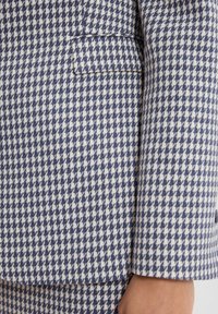 Tkanina w granatowo-białą kratę w typie houndstooth na dopasowanym żakiecie; posiada jedną kieszeń i gładką fakturę. Strukturalny design z wyraźnymi liniami.
