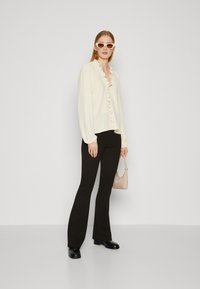 Cream blouse with ruffled collar, button front, and long puff sleeves paired with flared black trousers and a light brown handbag. Pink sunglasses.