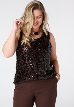 Femme blonde souriante regardant vers le bas, portant un haut sans manches noir à sequins, un pantalon marron, des colliers et bracelets superposés, une main dans la poche.
