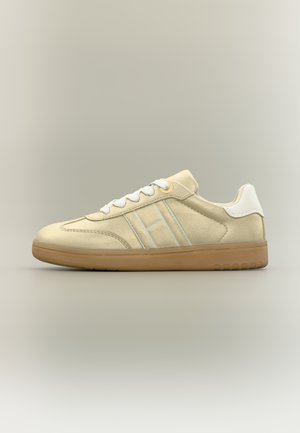 Scarpa da ginnastica beige con tomaia liscia in pelle scamosciata, dettagli bianchi, punta arrotondata e suola marrone chiaro. Presenta tre strisce diagonali sul lato.
