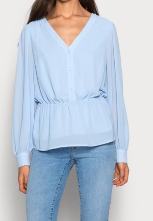 Femme portant un chemisier bleu clair à col en V avec manches longues, détails de boutons et taille froncée, associé à un jean bleu clair.