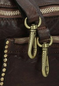 Detail van een bruin leren handtas met dubbele goudkleurige metalen clips, ritsaccenten en decoratieve gouden studs langs één kant. Textuurafwerking.