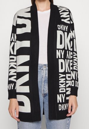 Personne portant un cardigan noir et blanc ouvert sur le devant avec le grand lettrage répété "DKNY" sur une chemise blanche et un jean bleu clair.