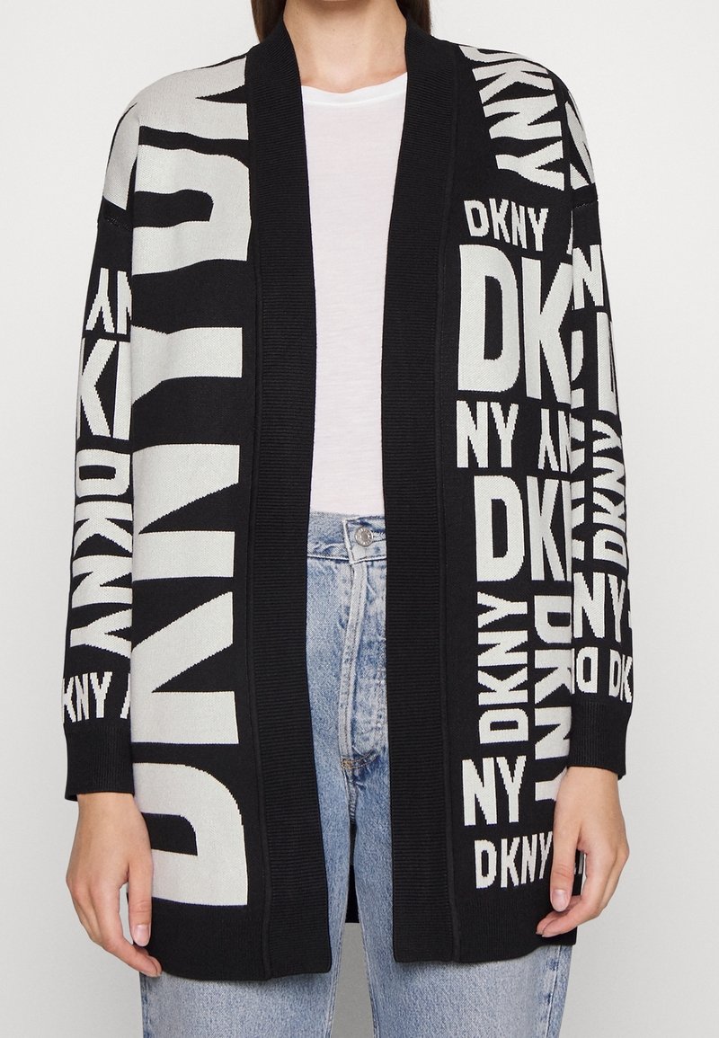 Personne portant un cardigan noir et blanc ouvert sur le devant avec le grand lettrage répété "DKNY" sur une chemise blanche et un jean bleu clair.