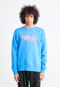 Blå Nike sweatshirt med rund hals, der har et stort pink logo og tekst på fronten. Lavet af blødt struktureret stof med lange ærmer.