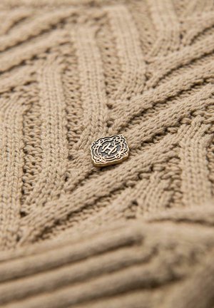 Beżowy sweter z dzianiny o teksturowanym wzorze w zygzak. Posiada dekoracyjny złoty emblemat z czarnym centralnym wzorem.