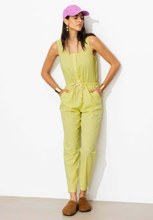 Femme portant une combinaison sans manches vert clair avec cordon, casquette rose, bracelets dorés et chaussures marron sans lacets, debout contre un mur blanc.