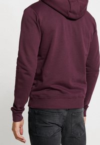 Person i en vinröd hoodie och svarta jeans, sedd bakifrån med vänsterhanden avslappnad vid sidan.