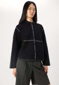 Zwarte textuurjacket met een staande kraag, witte stiksel details langs de naden en zwarte knopen. Draagt met grijze shorts.