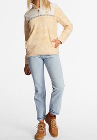 Lysebrun fleece-trøje med kontrasterende beige påskæring, flerfarvet vandret stribe og forlomme, kombineret med lyseblå jeans og brune støvler.
